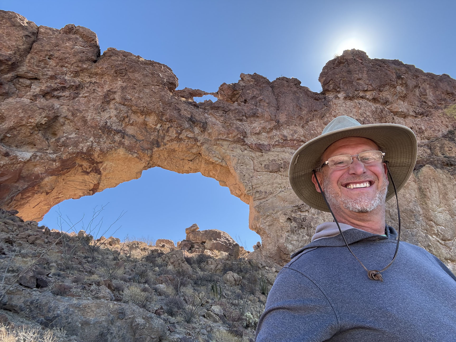 Shea in a sun hat and glasses stands outdoors near a large rocky arch formation. The sun shines brightly behind the rocks, creating a halo effect. The landscape is dry with sparse vegetation under a clear blue sky.