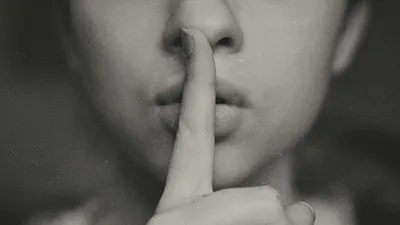 Close-up of a person’s face, slightly blurred, with their index finger held to their lips in a “shushing” gesture, suggesting silence or secrecy. The background is dark and out of focus.